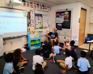 Children sitting in the floor in a classroom as their teacher (seated) reads aloud form a picture book she's holding up for them to see