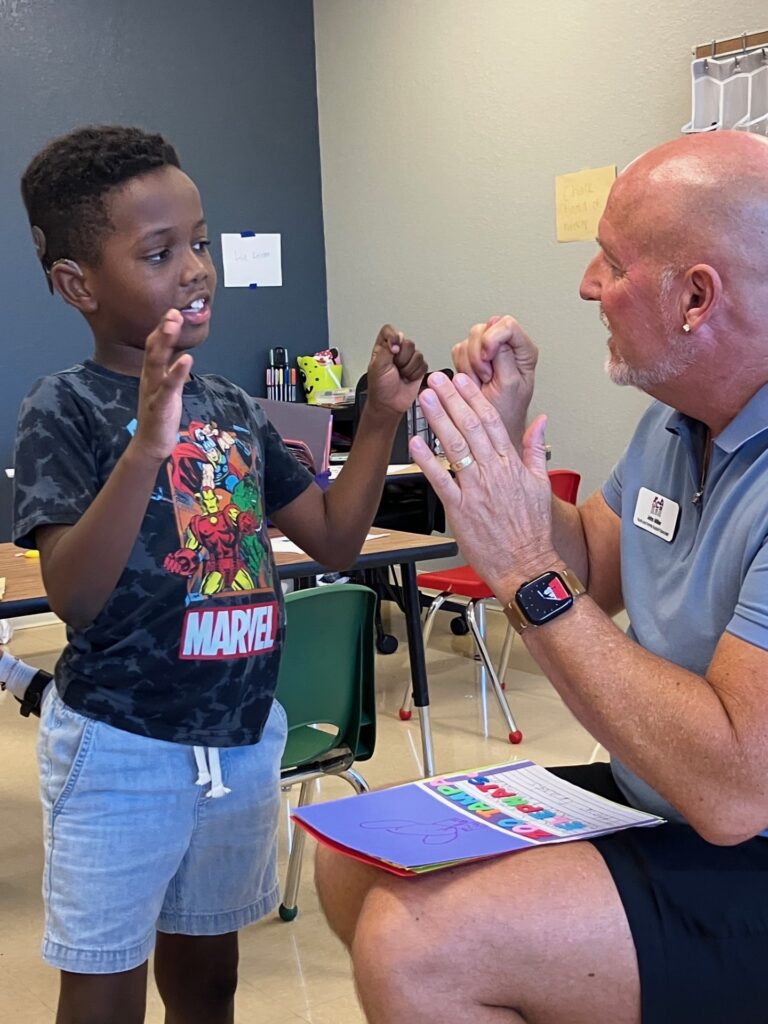 A standing boy wearing a hearing aid practices American Sign Language with a seated adult male in a classroom setting