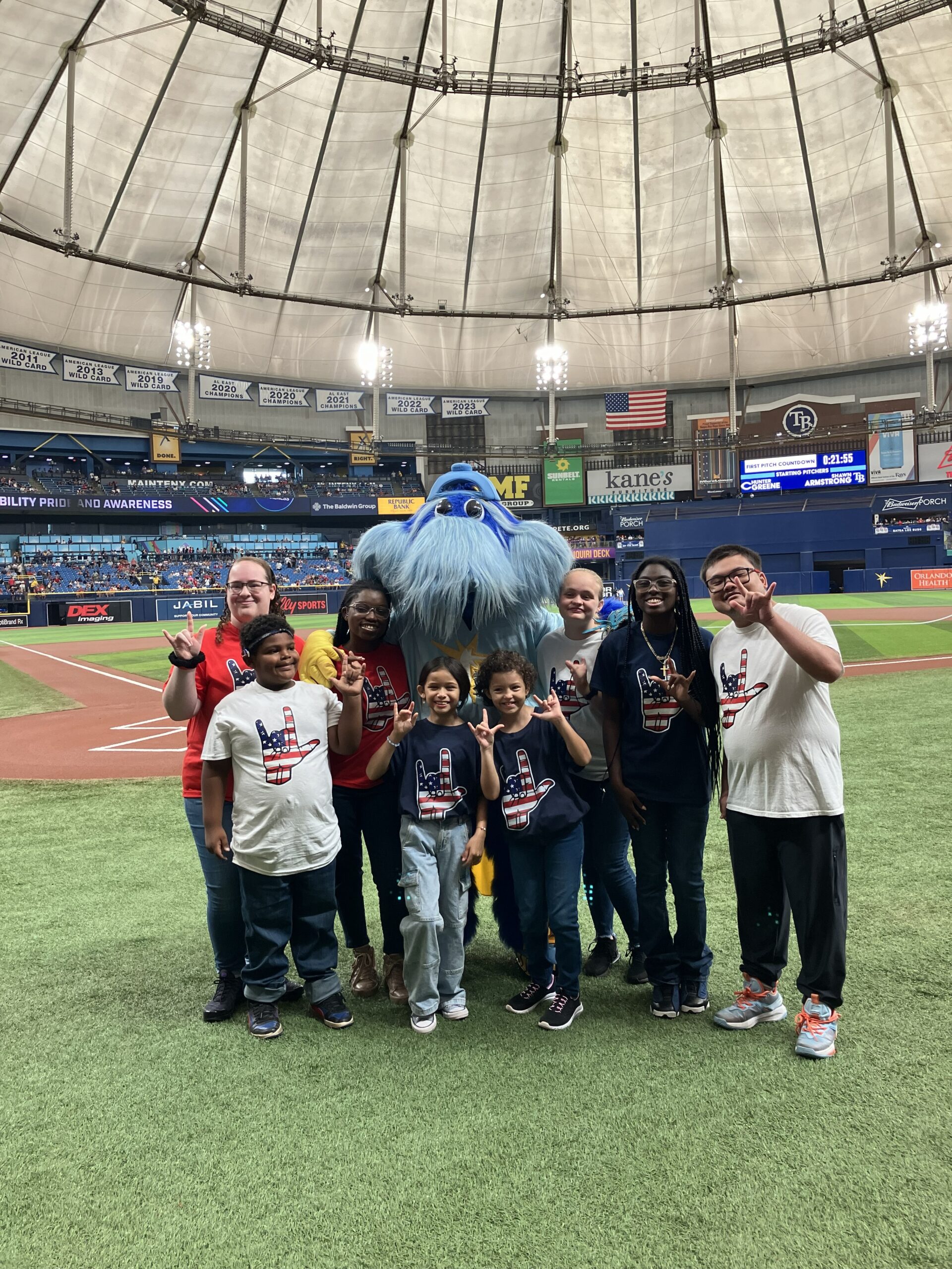 A group of adults and kids from FCD smiling in a group with the Rays team mascot on the field at the baseball stadium